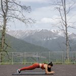 A woman doing a plank exercise on a mat outdoors with a scenic mountain view in the background.