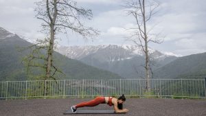 A woman doing a plank exercise on a mat outdoors with a scenic mountain view in the background.